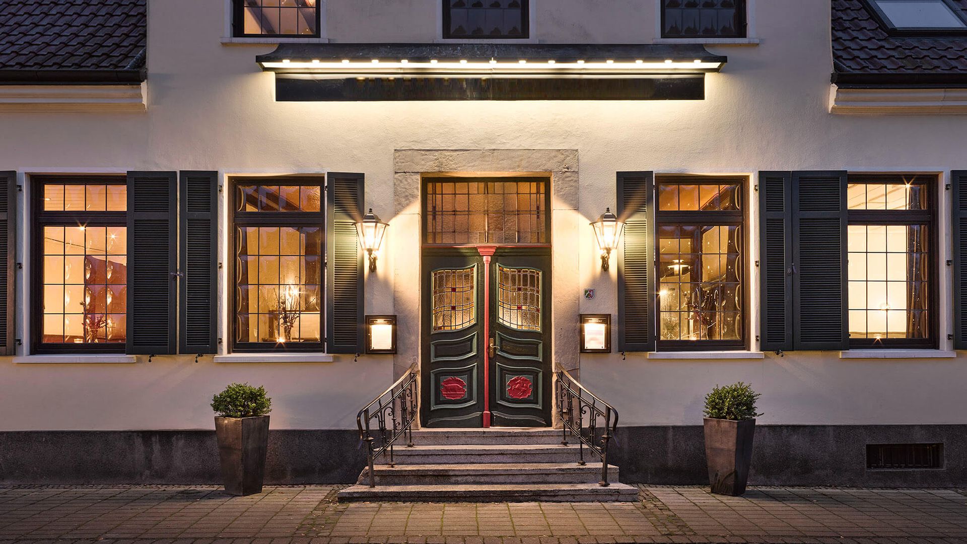 Elegantes Restaurant mit einer grünen Doppeltür mit Buntglas, warmer Beleuchtung und Topfpflanzen, eingebettet in eine charmante Fassade.