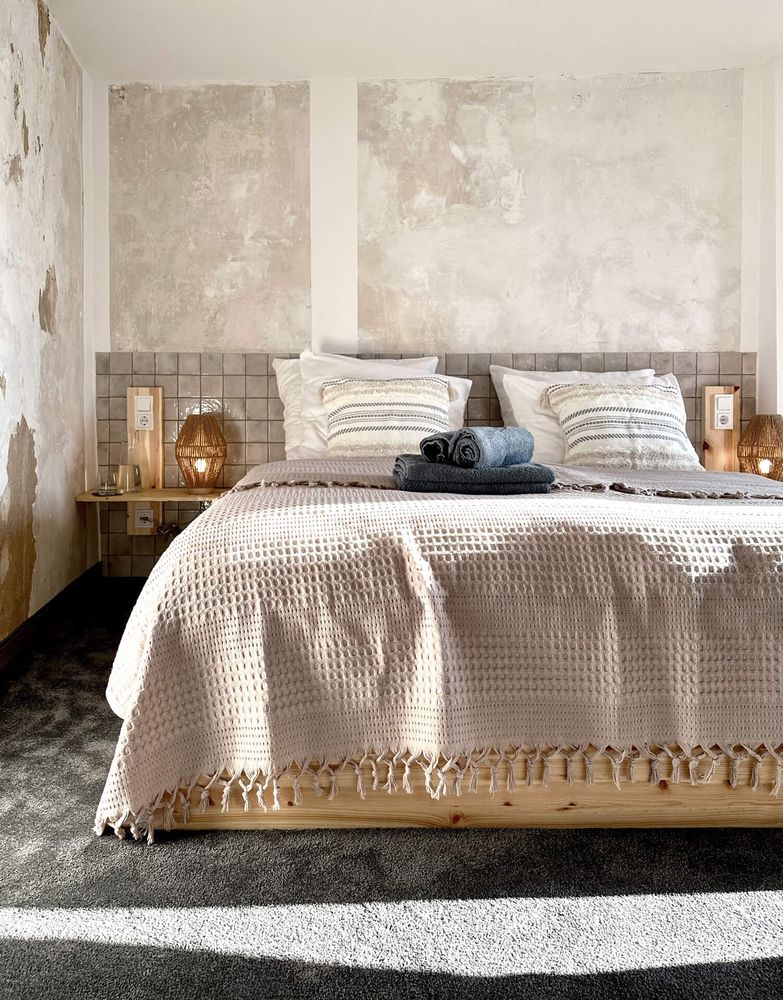 Gemütliches Schlafzimmer mit einem Holzbett mit einer strukturierten rosa Tagesdecke, weichen Kissen und warmer Umgebungsbeleuchtung vor verwitterten Wänden.